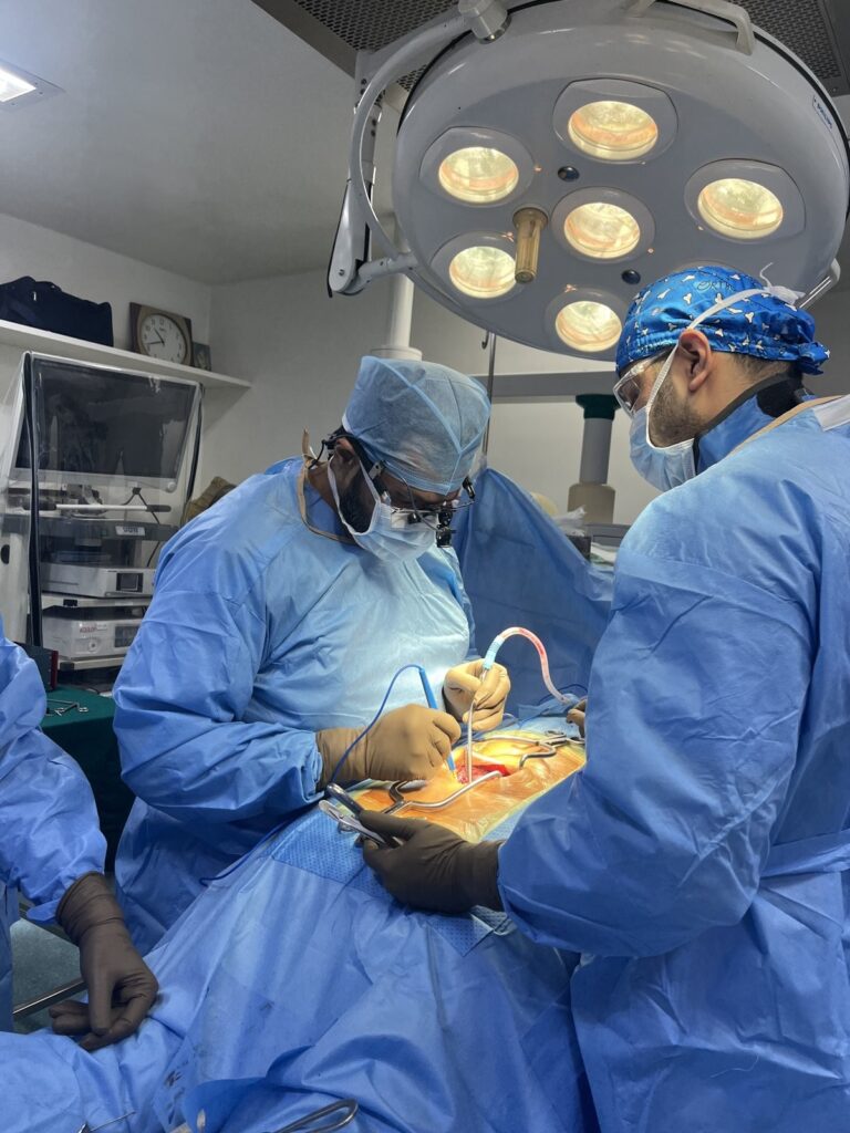 Spine surgeon wearing magnifying surgical loupes and a headlight operating under an overhead surgical light, assisted by a second surgeon in a blue gown