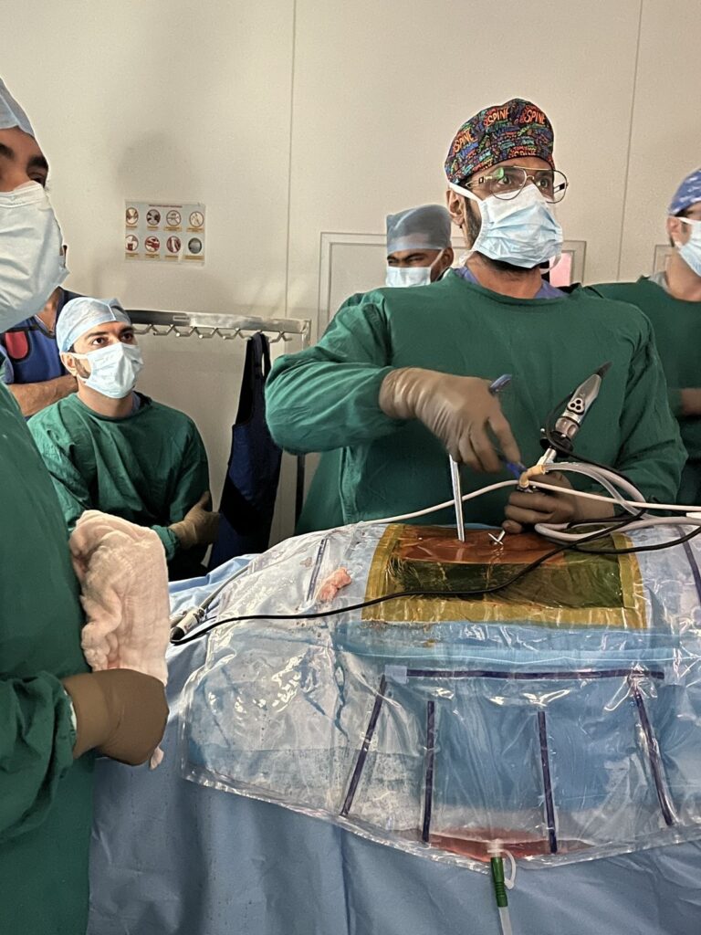 Spine surgeon in a green surgical gown and mask working on a draped patient's back during a spine procedure, with surgical team members watching in the operating theatre